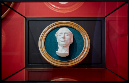 Eksponowana w Muzeum Książąt Czartoryskich jasna rzeźba przedstawiająca głowę mężczyzny ułożona na niebieskiej tkaninie otoczonej drewnianą owalną ramą.